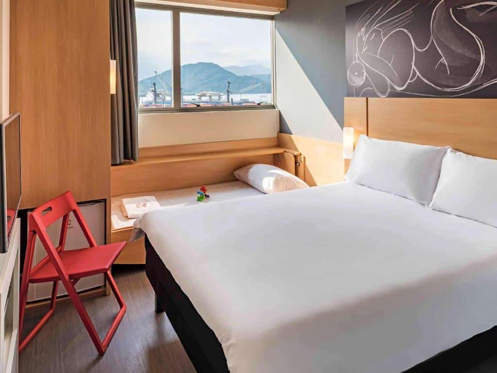 Quarto do ibis Santos Valongo com uma janela dando vista para o porto, uma cama de casal, um frigobar, uma televisão e uma cadeira