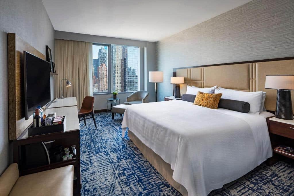 quarto do InterContinental New York Times Square, an IHG Hotel, com uma cama de casal à direita, carpete no chão, uma ampla mesa de trabalho em frente a cama com uma televisão presa à parede, e grandes janelas ao fundo do quarto.