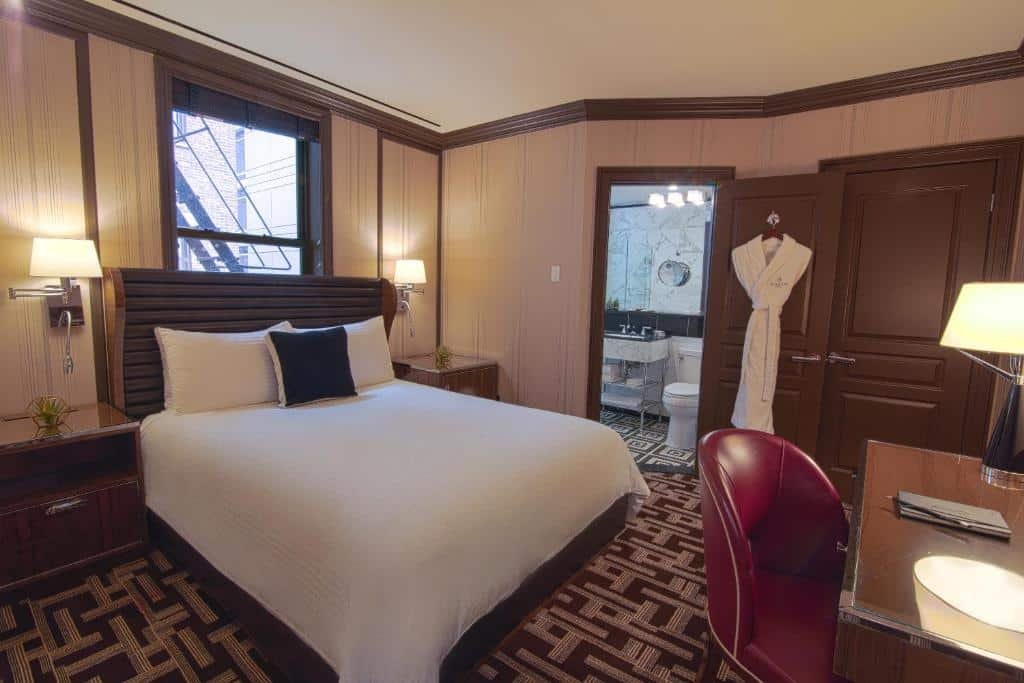 suíte do Iroquois New York Times Square hotel com uma cama de casal à esquerda, embaixo de uma janela que dá acesso a escada de incêndio. O quarto é coberto por carpete. Em frente a cama há uma mesa de madeira de trabalho com uma cadeira estofada vermelha. E ao fundo do quarto é possível ver uma porta aberta para o banehiro.