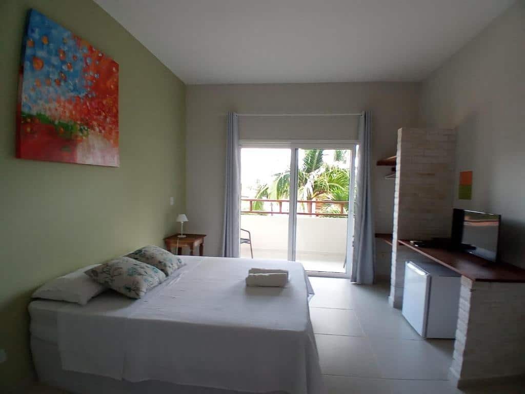 Quarto do Kauano Pousada Barequeçaba com cama de casal, cômoda de madeira do lado esquerdo com luminária, em frente a cama frigobar com TV.