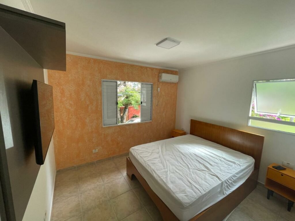 Quarto do Lar Doce Lar Toque Toque Grande com cama de casal do lado direito da imagem com uma cômoda em cada lado com uma TV a frente presa na parede. Representa airbnb na praia de Calhetas.