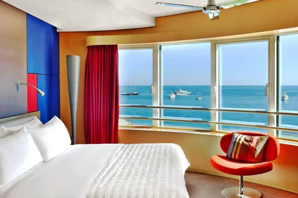 Quarto no Le Méridien Beach Plaza. Uma cama de casal no lado esquerdo, de frente uma poltrona. No lado direito uma paredes de vidro com vista para o mar. Foto para ilustrar post sobre Hotéis em Mônaco.