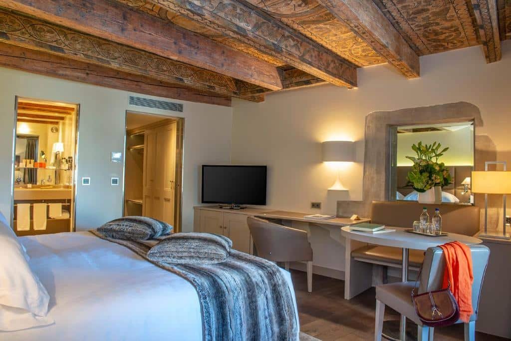 Foto de um quarto do Les Armures, para ilustrar o post de hotéis em Genebra. Uma cama de casal está no canto inferior esquerdo da foto, de costas para a imagem. Na frente da cama, há uma escrivaninha com TV em cima, abajur e duas cadeiras. Um espelho também está fixo na parede. No lado esquerdo do quarto, do lado da escrivaninha está a porta da suíte e outra porta para o banheiro.