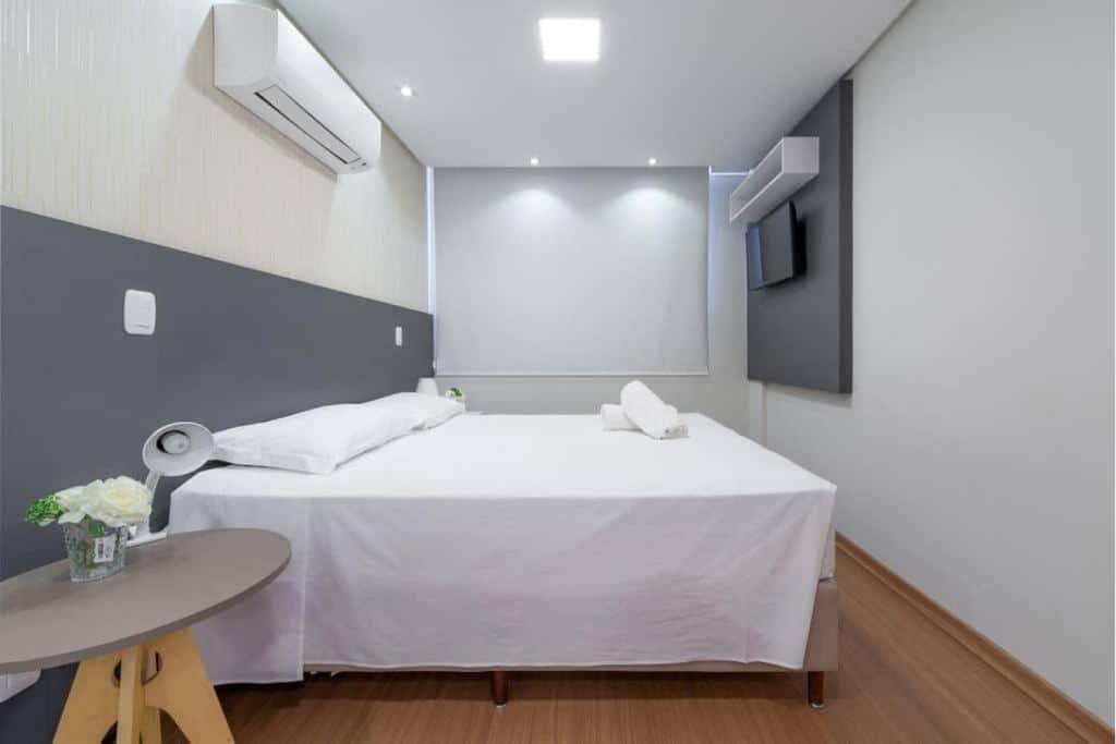 Quarto no Lindo Apto De 2 Quartos Em Gramado - Ed Paradiso, uma janela com persiana, uma televisão, um ar-condicionado, uma cama de casal, um pequena mesinha com um abajur, ambiente em tons de branco e cinza, para representar airbnb em Gramado