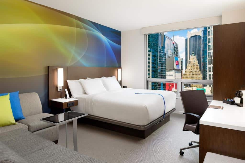 quarto do LUMA Hotel perto da Times Square com um sofá de canto de 5 lugares localizado no canto inferior esquerdo, ao lado esquerdo da cama de casal. No lado direito da cama há uma janela ampla com vista para a Times Square e uma mesa de trabalho em frente a cama.
