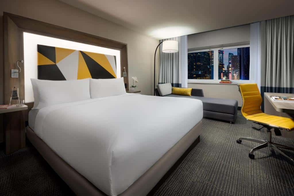 quarto do M Social Hotel Times Square New York com uma cama de casal à esquerda da imagem, ao lado de um sofá divã abaixo da janela, ao fundo do quarto. Na outra extremidade da cama há uma mesa com cadeira estofada amarela e giratória.
