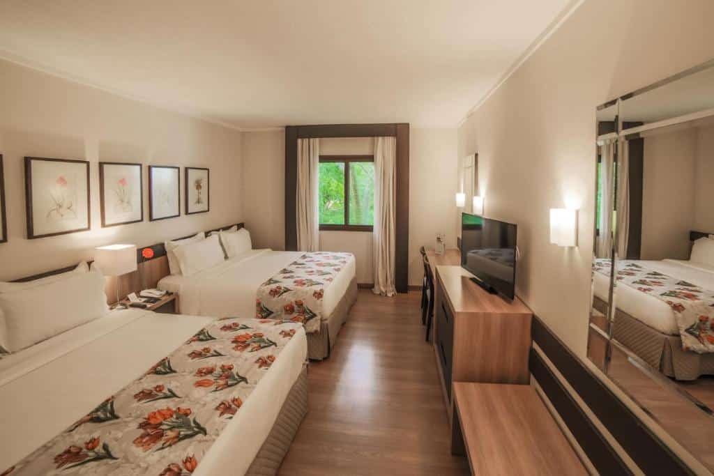 Quarto do Mabu Thermas Grand Resort com duas camas de casal separadas por uma mesinha de cabeceira. Em frente as camas tem um espelho e alguns móveis com uma tv em cima. Do lado da cama esquerda tem uma janela de vidro com cortinas.
