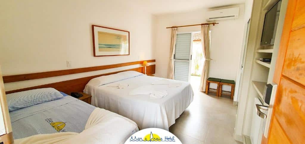 Quarto do Mar e Praia Hotel, duas camas de casal, uma sacada, um quadro sob a cama, uma televisão e alguns armários, além de um frigobar, para representar pousadas na Praia da Enseada em Ubatuba