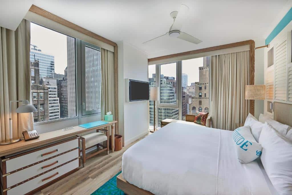 suíte do Margaritaville Resort Times Square hotel com uma cama de casal à direita com vista para a cidade. No lado esquerdo da cama há janelas panorâmicas cobrindo toda a parede mostrando diversos prédios de Manhattan.