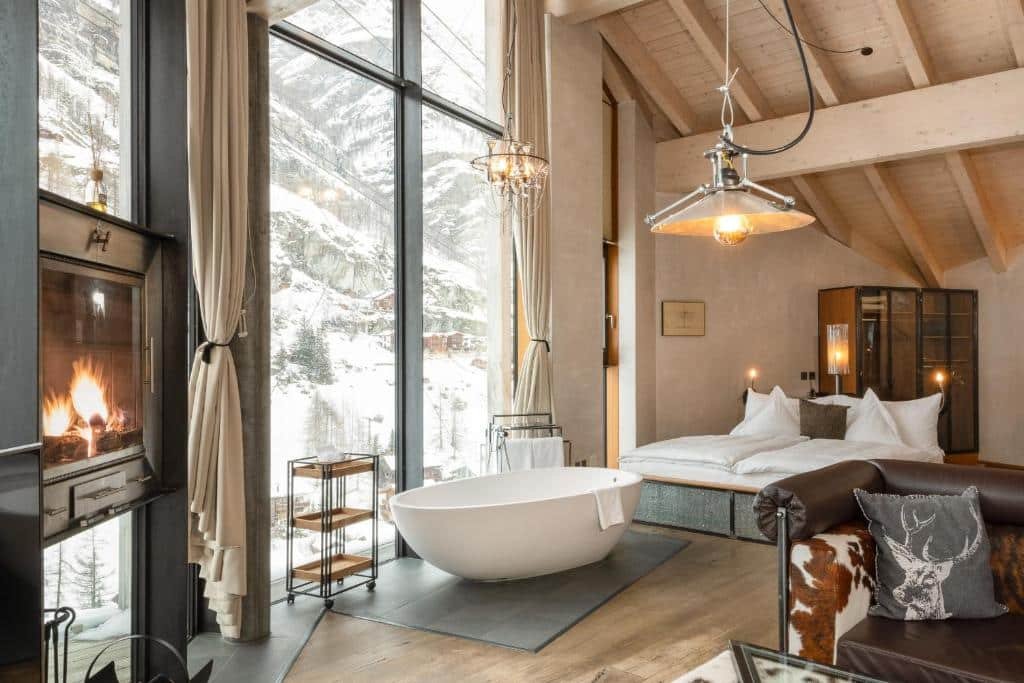 Quarto do Matterhorn FOCUS Design Hotel ao fundo uma cama de casal, em frente a cama do lado esquerdo banheira de hidromassagem em frente a uma janela ampla com vista para as montanhas e em frente a banheira do lado direito da imagem uma lareira. Representa onde ficar em Zermatt.