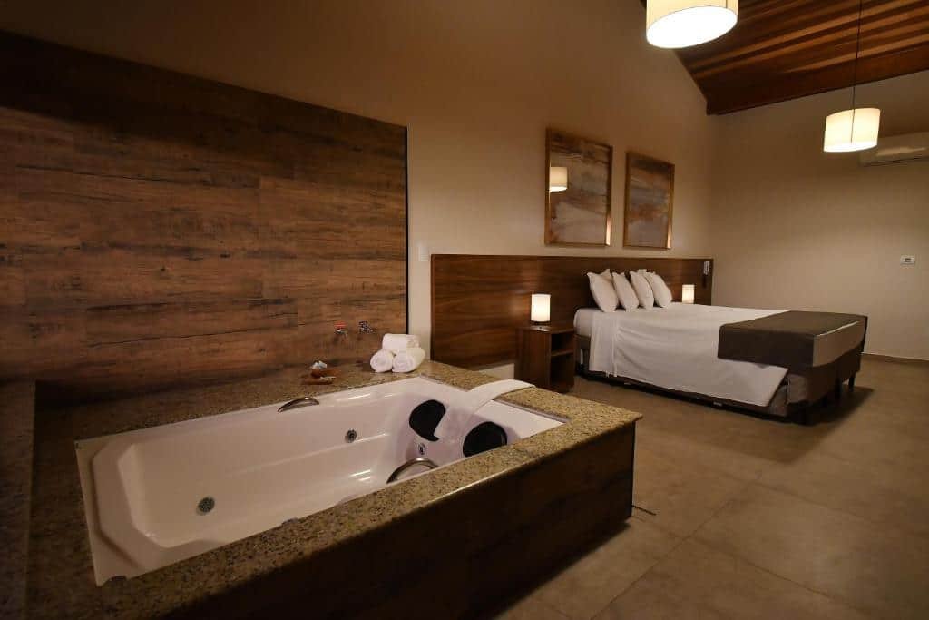 Quarto amplo na Molise Hotel Fazenda com uma banheira, uma cama de casal, uma cabeceira com luminárias e dois quadros sob a cama, para representar hotéis em Serra Negra