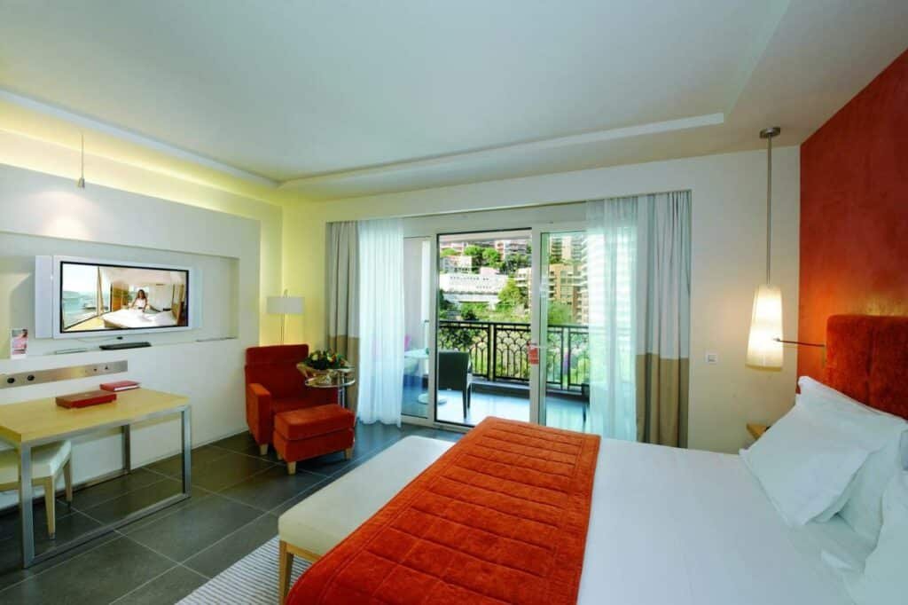 Quarto do Monte-Carlo Bay Hotel & Resort. Uma cama de casal no lado direito, de frente uma mesa com uma cadeira, uma poltrona uma televisão. No fundo, a sacada do quarto. Foto para ilustrar post sobre Hotéis em Mônaco.
