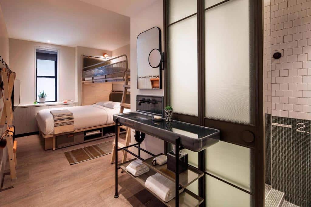quarto do MOXY NYC Times Square com um banheiro na entrada do quarto, no lado direito, com uma pia retrô do lado de fora, e um box e vaso sanitário separados por uma porta de vidro. Mais ao fundo do quarto, no lado esquerdo, há uma cama de casal com uma cama de solteiro acima, como uma beliche de três lugares