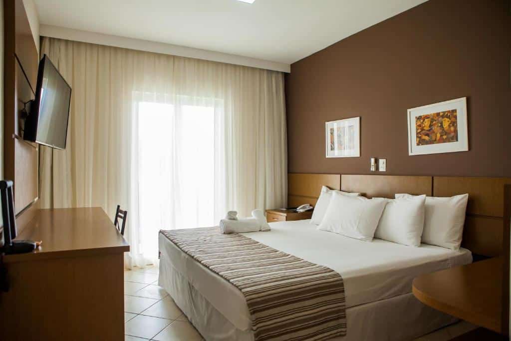 Quarto do Mundial Parque Hotel. A cama está centralizada, no lado direito é possível ver um pedaço da mesa, no lado esquerdo da cama há uma pequena cômoda e na frente há outra cômoda e uma TV. Imagem para ilustrar o post hotéis em Viçosa.