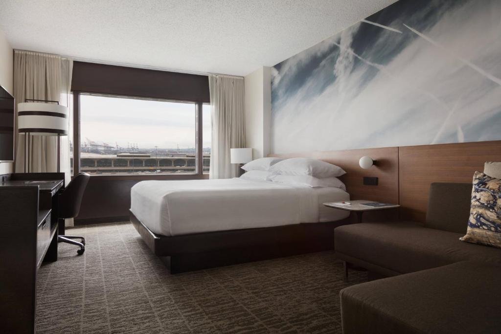Imagem de um quarto confortável e aconchegante no Newark Liberty International Airport Marriott. O quarto possui uma decoração moderna com cores suaves e móveis elegantes. Há uma cama espaçosa e macia no centro do quarto, com lençóis limpos e bem arrumados. Nas laterais da cama, há mesas de cabeceira com luminárias de leitura. À direita, encontra-se uma escrivaninha bem iluminada, equipada com uma cadeira ergonômica para trabalho ou estudo. A parede oposta à cama possui uma grande TV de tela plana, perfeita para entretenimento. Há também um armário espaçoso para guardar pertences pessoais. O quarto proporciona uma atmosfera tranquila e agradável para descanso e relaxamento.