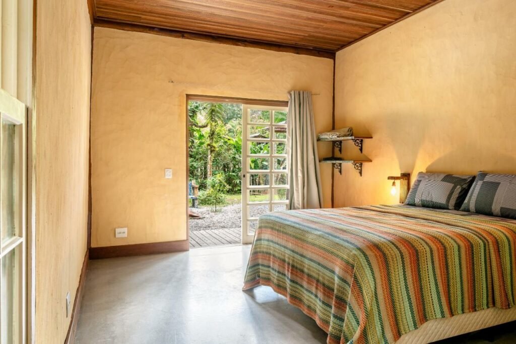 Quarto do Nosso quintal é a praia de Maresias com cama de casal do lado direito e do lado direito uma porta que dá acesso a uma porta.