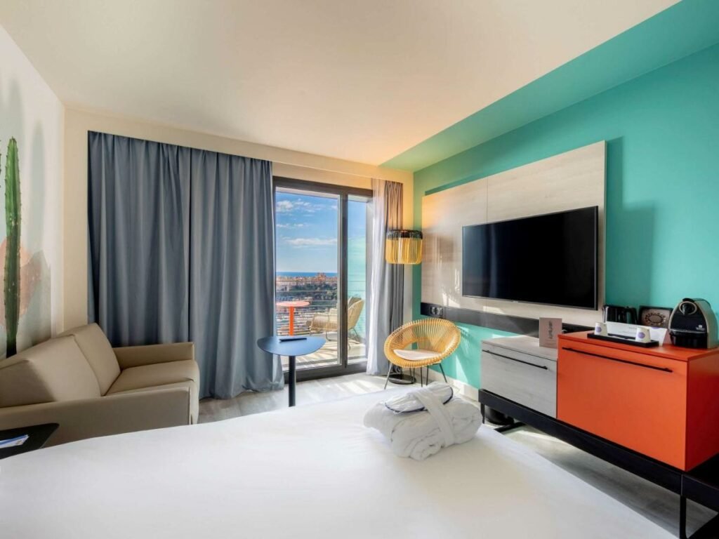 Quarto do Novotel Monte-Carlo. Uma cama de casal no lado esquerdo com uma poltrona. De frente, um pequeno armário, uma televisão e uma cadeira. No fundo do quarto a varanda. Foto para ilustrar post sobre Hotéis em Mônaco.