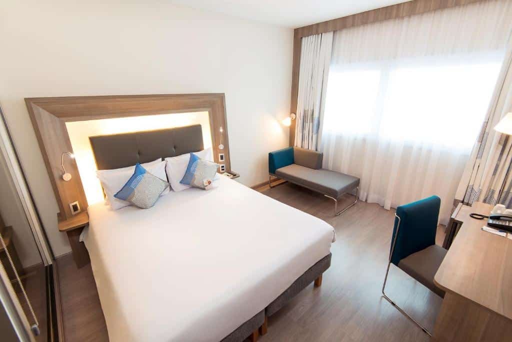 Quarto do Novotel Santos Gonzaga com uma cama de casal, um pequeno sofá uma janela com cortinas e uma mesa de escritório com uma cadeira