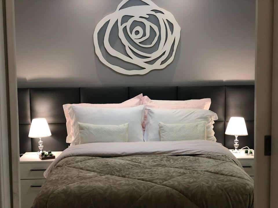 Quarto no Ocean Flat Premium, com uma cama de casal cheia de travesseiros, tudo em detalhes de prata e cinza, um quadro desenhando uma flor sob a cama de forma minimalista, e dois abajures (um de cada lado da cama)