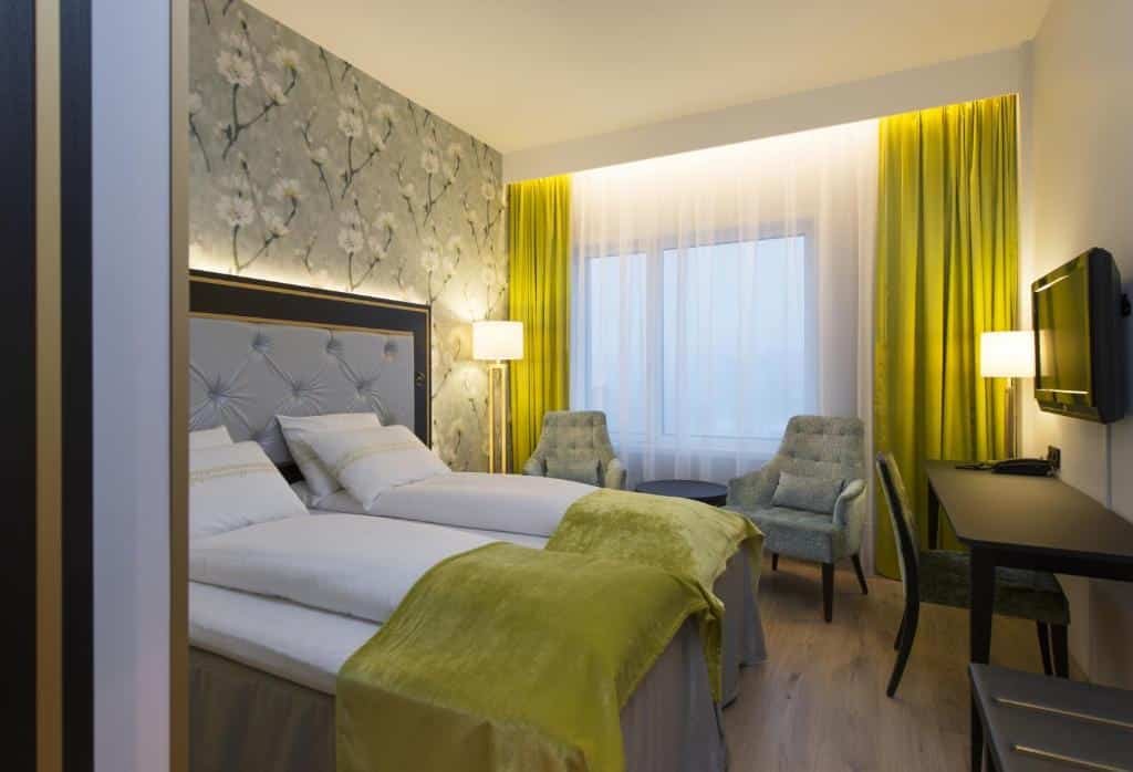 Quarto do Thon Hotel Opera com duas camas de solteiro, duas poltronas cinzas do lado esquerdo perto da janela. Mesa de trabalho em frente as camas com TV na parede.