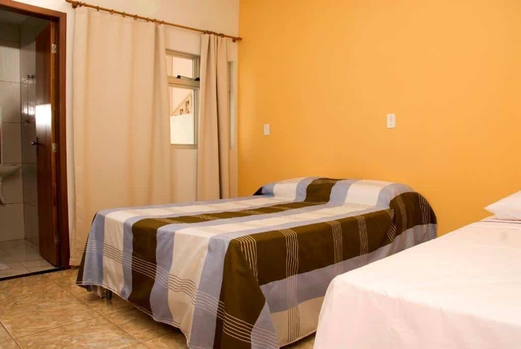 Quarto do Orquídea Hotel. No lado esquerdo está a porta para o banheiro, uma pequena janela e uma cama de casal. No lado direito é possível ver parte de uma cama de solteiro. Imagem para ilustrar o post hotéis em Viçosa.