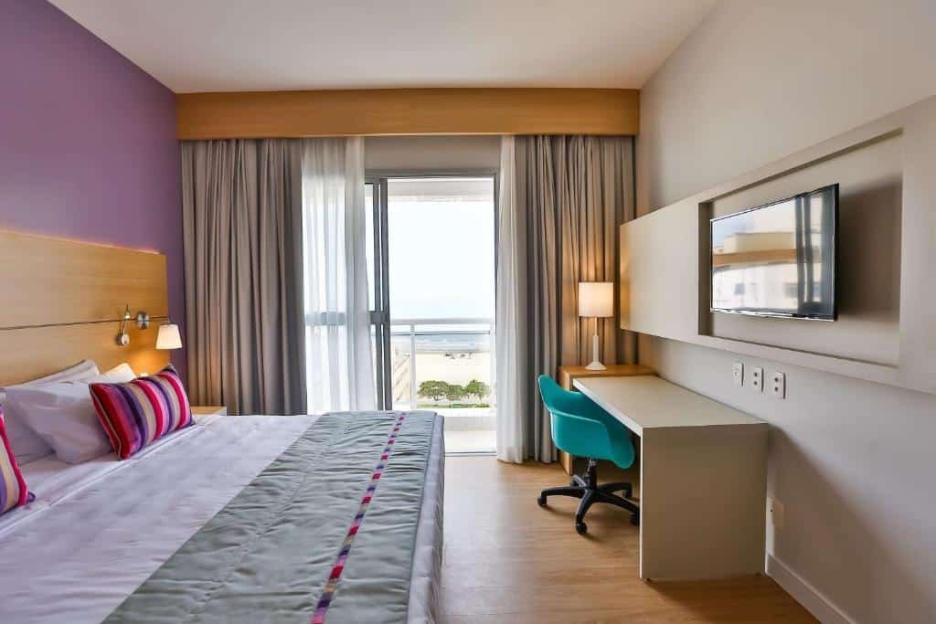 Quarto do Park Inn by Radisson Santos com uma varanda dando vista para o mar, chão de madeira, cama de casal, uma televisão e uma bancada com uma cadeira giratória