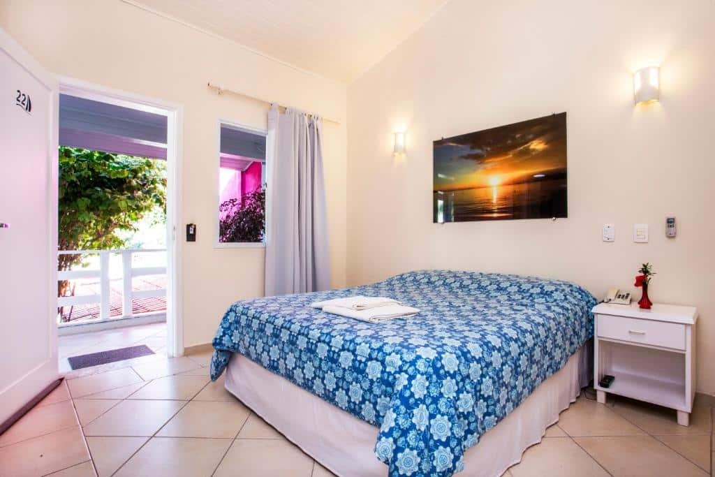 Quarto da Pousada Azul da Cor do Mar com cama de casal na área central, cômoda branca do lado direito, quadro com paisagem de pôr-do-sol na praia e do lado esquerdo porta tem uma janela com cortinas brancas.