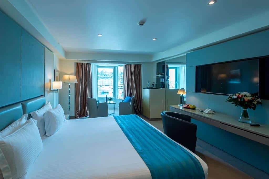 Quarto do Port Palace. Uma cama de casal no lado esquerdo e de frente uma televisão, com uma mesinha e cadeira. No fundo, uma mesa redonda com duas cdeiras e a vista do quarto para o porto. Foto para ilustrar post sobre Hotéis em Mônaco.