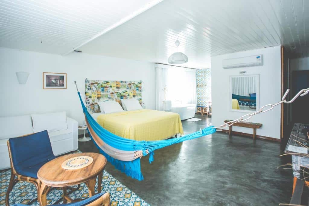 Quarto da Pousada a Capela. Uma mesa redonda com duas cadeiras e um sofá atrás no lado esquerdo. No meio uma rede, no lado direito uma cama de casal, uma banheira, uma janela um espelho, um banco e um ar-condicionado. Foto para ilustrar post sobre pousadas em Arembepe.