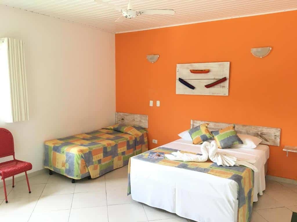 Quarto em Pousada Baía das Conchas com cama de casal e uma de solteiro, local amplo, roupa e cama e decoração em laranja claro e azul, uma cadeira, uma janela, para representar pousadas na Praia da Enseada em Ubatuba