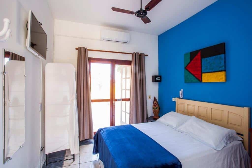 Quarto da Pousada Azul Banana com cama de casal ao meio, TV a frente da cama colocada na parede do lado direito de um espelho, do lado esquerdo portas de madeiras com cortinas marrom que dá acesso a varanda.