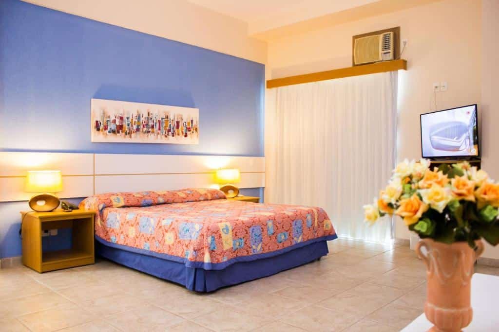 Quarto da Pousada Barlavento. Um vaso na frente, no canto direito, atrás uma televisão, ar-condiconado e a janela do quarto. No canto esquerdo, uma cama de casal no meio e um criado mudo de cada lado com um abajur. Foto para ilustrar post de pousadas em Iriri (ES).