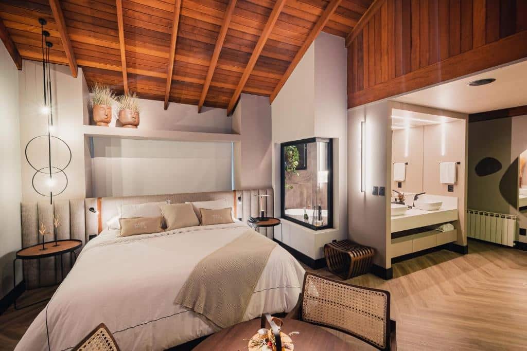 Quarto da Pousada Bella Terra, um local super amplo, com arquitetura minimalista e moderna, uma cama de casal, um banheiro com banheira, lustres, decoração simples, uma mesa com dois lugares, toalhas e travesseiros sob a cama