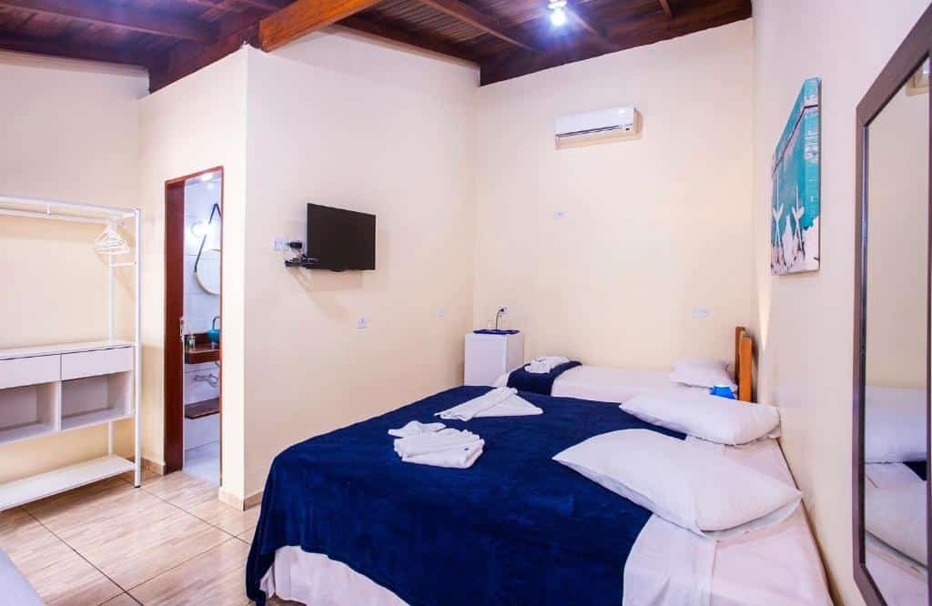 Quarto da Pousada Villa Blu Maresias com cama de casal do lado direito e do lado esquerdo cama de solteiro; em frente as camas tem uma TV, do lado esquerdo perto da cama, uma de solteiro e um frigobar.