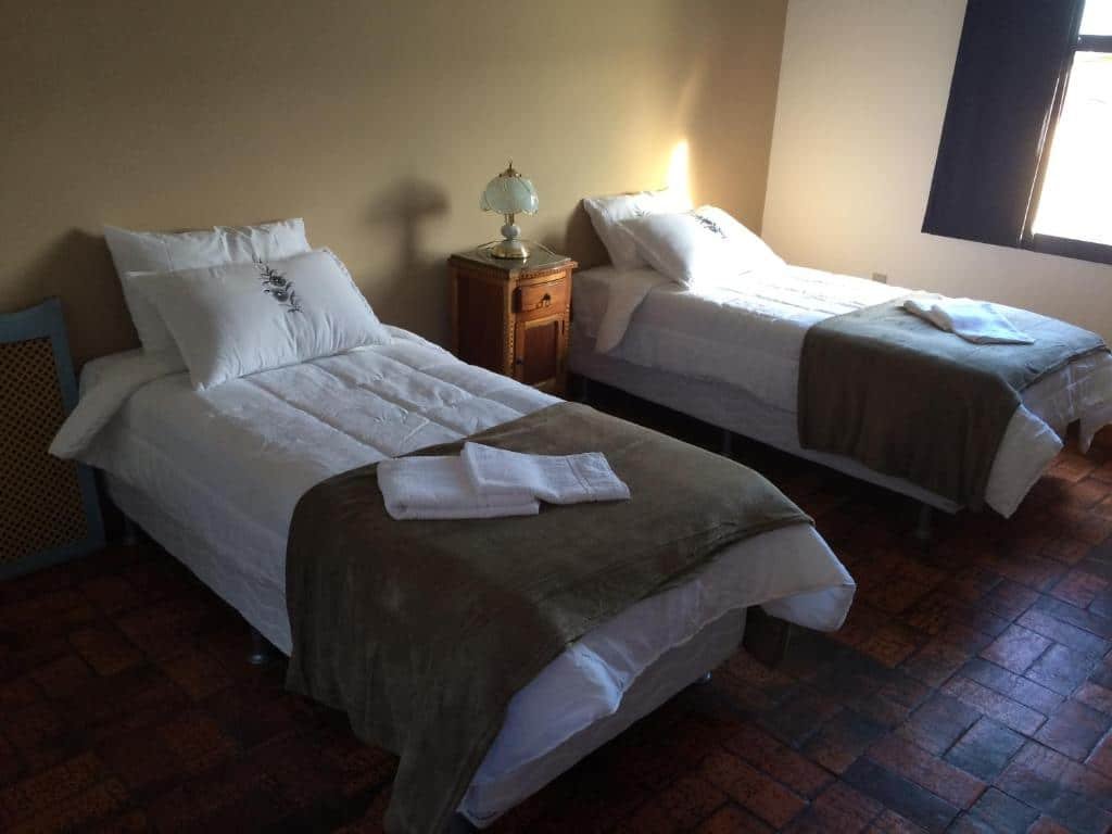 quarto da Pousada Casarão da Pedra em Congonhas com duas camas de solteiro e uma cômoda entre as duas. Há um abajur antigo e delicado em cima do móvel