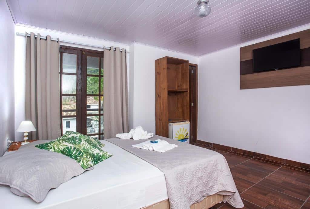 Quarto da Pousada Castelinho com cama de casal, do lado esquerdo com luminária e uma cômoda dom frigobar.