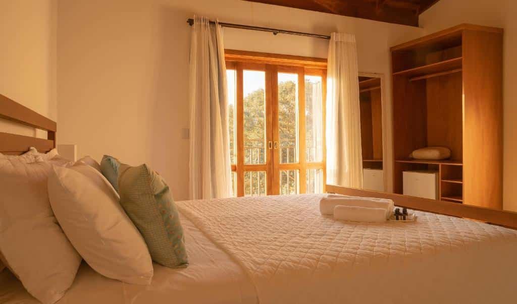 Quarto na Pousada Cheiro da Terra, uma cama de casal, uma sacada com vista para a grama, um espelho, um armário de conceito aberto, toalhas, almofadas e travesseiros sob a cama, para representar pousadas em Escarpas do Lago