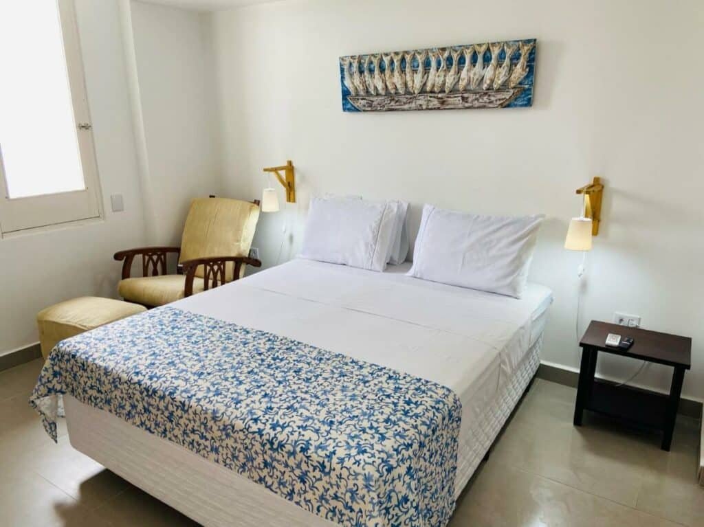 Quarto na Pousada Cocal da Praia com uma cama de casal, uma janela, dois pequenos abajures, uma poltrona, e um quadro com decoração praiana, tudo em azul e branco