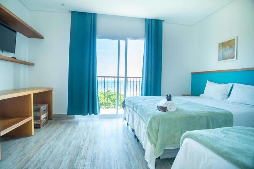Quarto de hotel com duas camas, detalhes em azul-petróleo, TV, prateleiras e obras de arte na parede. Grande janela com cortinas azul-petróleo com vista para o mar.
