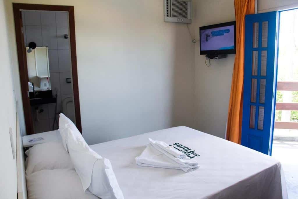 Quarto da Pousada das Flores. Uma cama de casal na frente, atrás, no lado esquerdo a porta do banheiro. De frente para a cama uma televisão e a porta da sacada do quarto.
