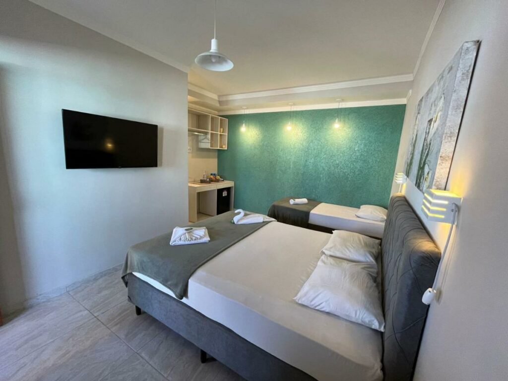 Quarto da Pousada Fast Inn com uma cama de casal, uma de solteiro, uma pequena bancada, um frigobar, duas luminárias e uma televisão