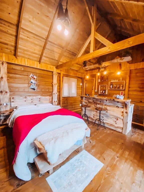 Quarto da Pousada Flocos de Neve, um dos chalés em Urubici, com estrutura totalmente em madeira, incluindo a cabaceira da cama, a bancada e um banco aos pés da cama. Há duas cadeiras altas, luminárias de teto e outras decorações.