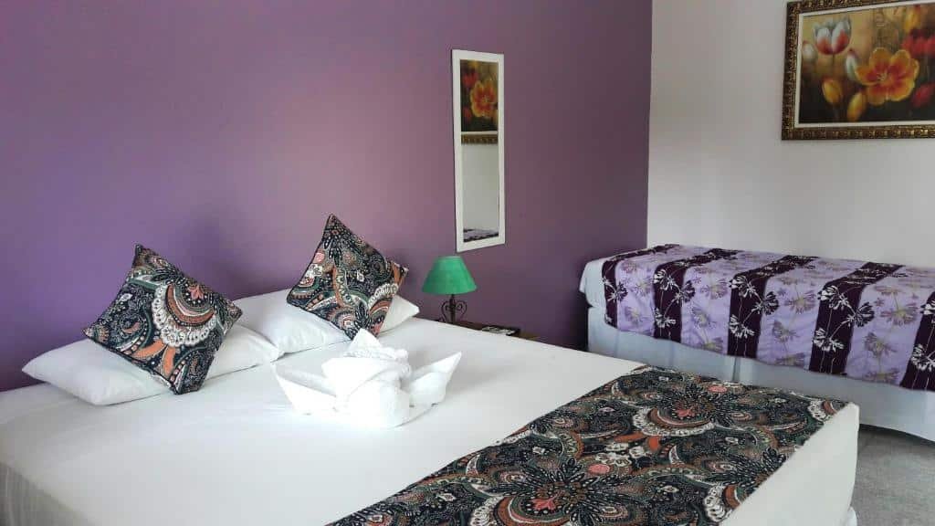 Quarto Pousada Flores do Lázaro, parede roxa claro, uma cama de casal com roupa de cama desenhada, uma cama de solteiro, um espelho, uma abajur e um quadro