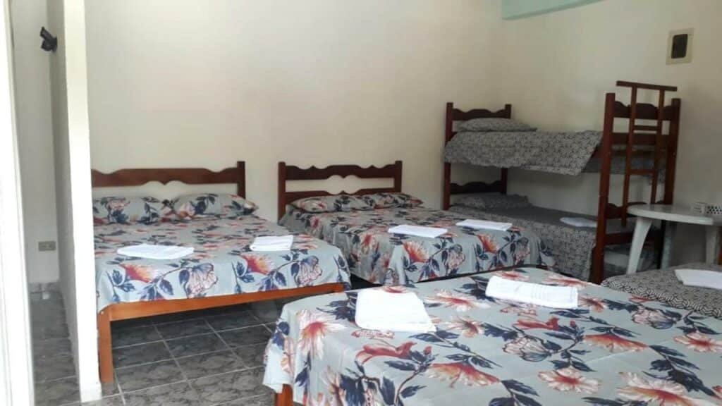 Quarto da Pousada Geres Peruíbe. Uma cama de casal de frente pra duas camas de casais. No fundo, do lado das duas camas, no canto direito, uma cama beliche e de frente pra cama uma mesa de plástico.