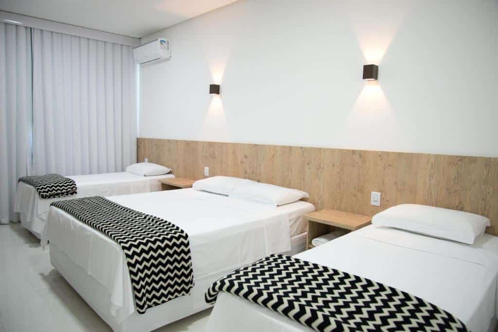 Quarto na Pousada Kanto da Ilha Suites, uma cama de casal, duas de solteiro, iluminação indireta, duas mesinhas de cabeceira, uma varanda com cortinas, travesseiros e cobertas preta e branca sob as acomodações, para representar pousadas em Escarpas do Lago