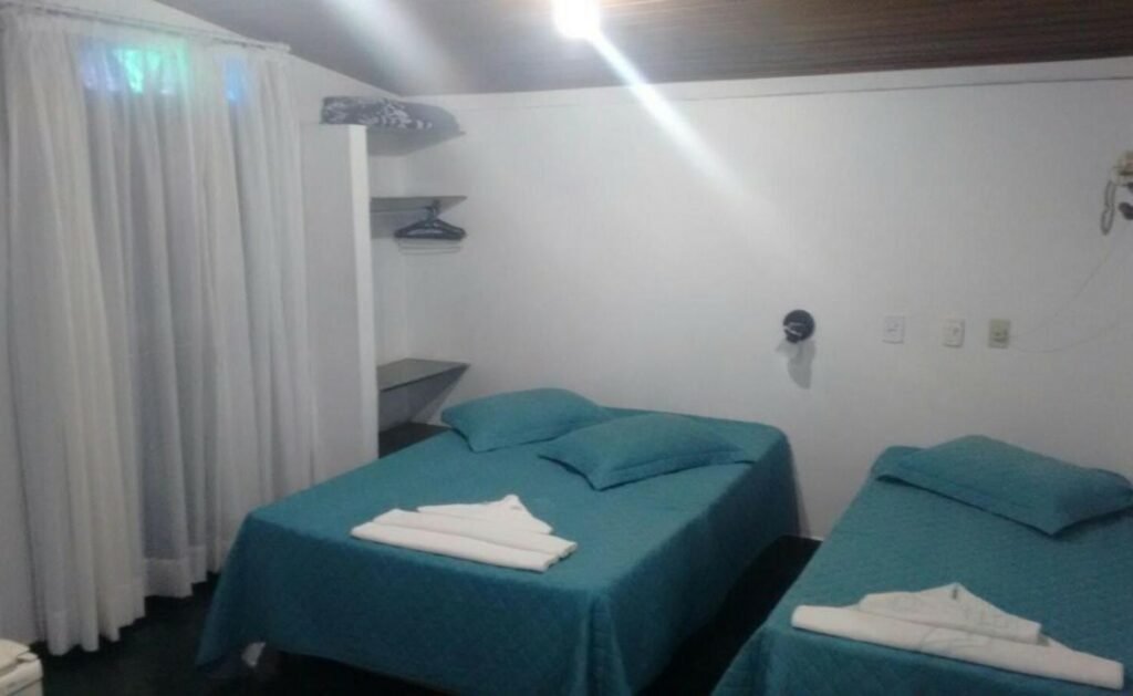 Quarto da Pousada da Memeia. Uma cama de solteiro no lado direito e no meio uma cama de casal. No canto esquerdo, a cortina do quarto e no fundo umas prateleiras de apoio. Foto para ilustrar post de pousadas em Iriri (ES).