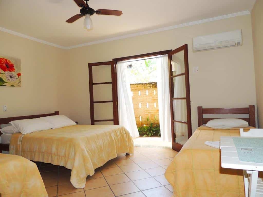 Quarto da Pousada Mosaico Brasil com cama de casal do lado esquerdo, uma cama de solteiro ao lado e do lado direito outra cama de solteiro; na área central portas de madeira que dá acesso a um jardim.