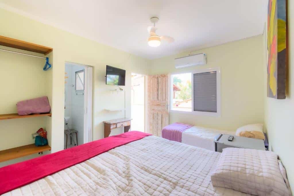Quarto na Pousada Movimenthai. Uma cama de casal, um frigobar e uma cama de solteiro. Do lado da cama de solteiro uma janela e em cima um ar-condicionado. De frente para as camas três prateleiras, a porta do banheiro, uma televisão e a porta do quarto. Foto para ilustrar post sobre Pousadas em Boracéia.