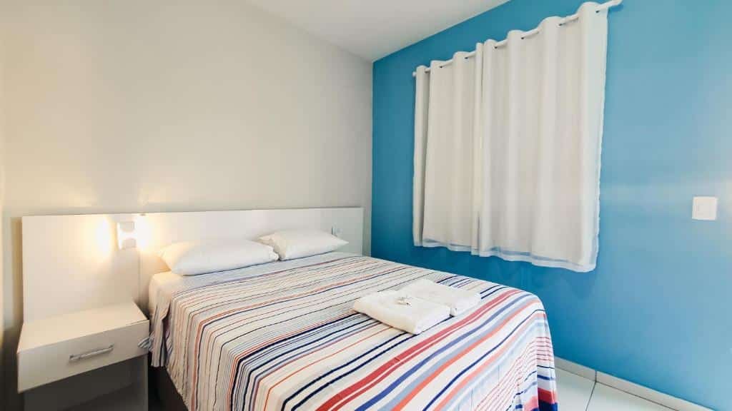 Quarto da Pousada Paraiso de Arempebe. Uma cama de casal no meio, de cada lado uma gaveta e uma luminária. No lado esquerdo da cama uma janela com cortina. Foto para ilustrar post sobre pousadas em Arembepe.