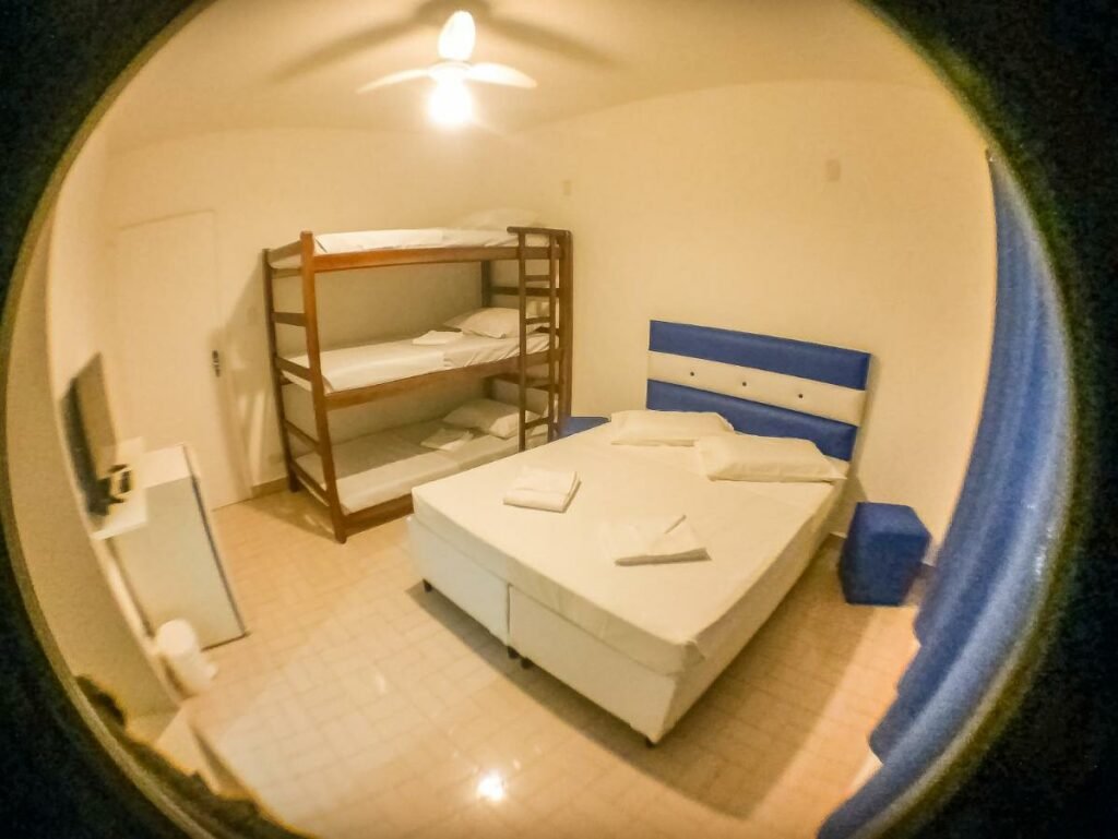 Quarto da Pousada Peruibe 455. Uma cama de casal e atrás uma cama beliche. De frente pra cama de casal um frigobar e uma televisão.